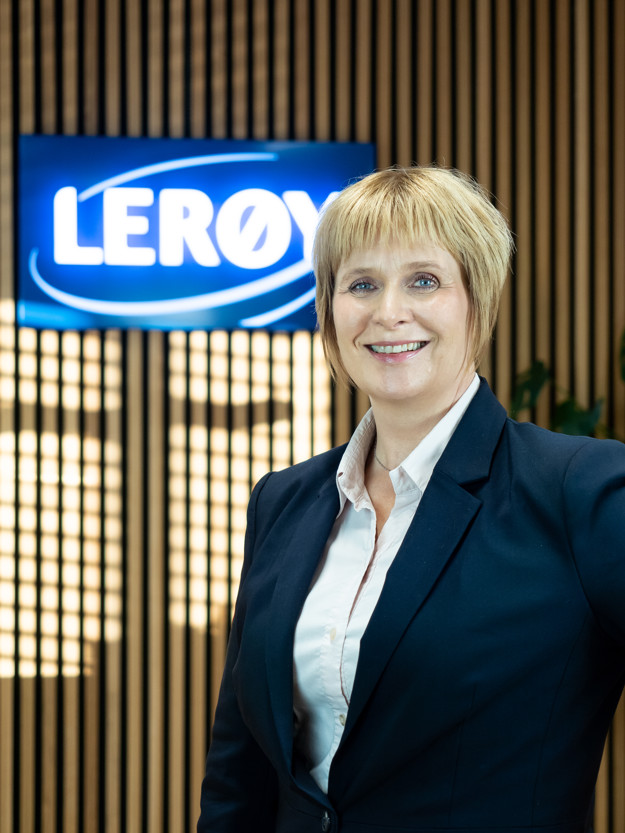 A smiling woman with a white shirt and blue blazer, behind her is a bright  "Lerøy" sign hanging on the wooden wall.